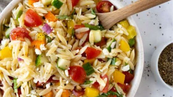 Orzo salad with peppers and tomatoes in white bowl with wooden spoon