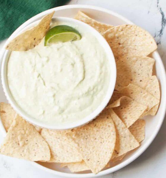 Avocado dip in white bowl on white plate with tortilla chips