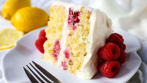 A slice of lemon raspberry cake on a white plate with raspberries on the side and lemons in the background