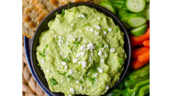 Avocado dip with feta cheese in a black bowl with crackers, cucumbers, carrots and celery on the side