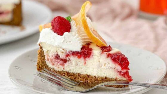 Lemon Raspberry cheesecake with raspberries and a slice of lemon on top sitting on a white dessert plate with a fork.