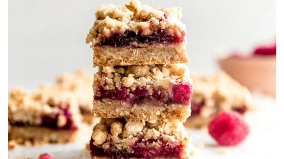 Raspberry oatmeal bars stacked on white surface with raspberry next to them.