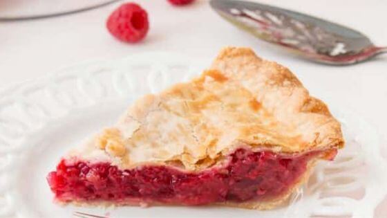 A piece of raspberry pie on a white scalloped plate with one red raspberry next to it.