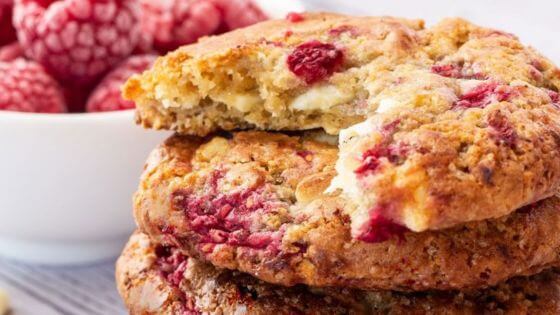 Raspberry and white chocolate cookies stacked on a striped towel with a white bowl of raspberries next to them.
