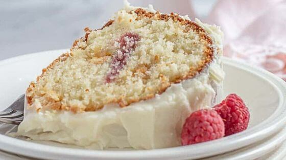 A slice of a raspberry bundt cake with white chocolate on a white plate with raspberries on the side.