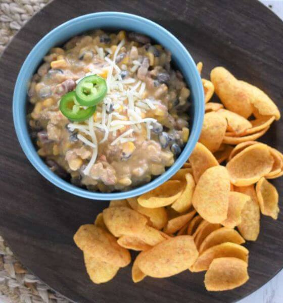 Slow cooker chili cheese dip in blue bowl sitting on a black plate with corn chips