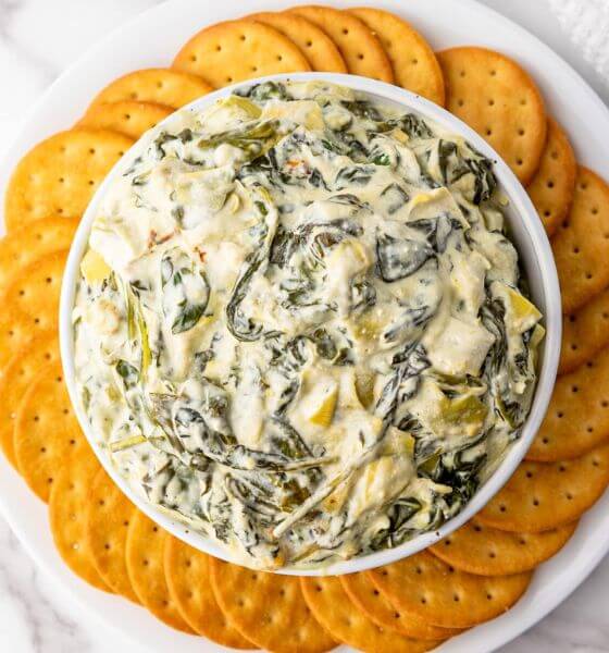 Spinach artichoke dip in white bowl with round crackers circling it